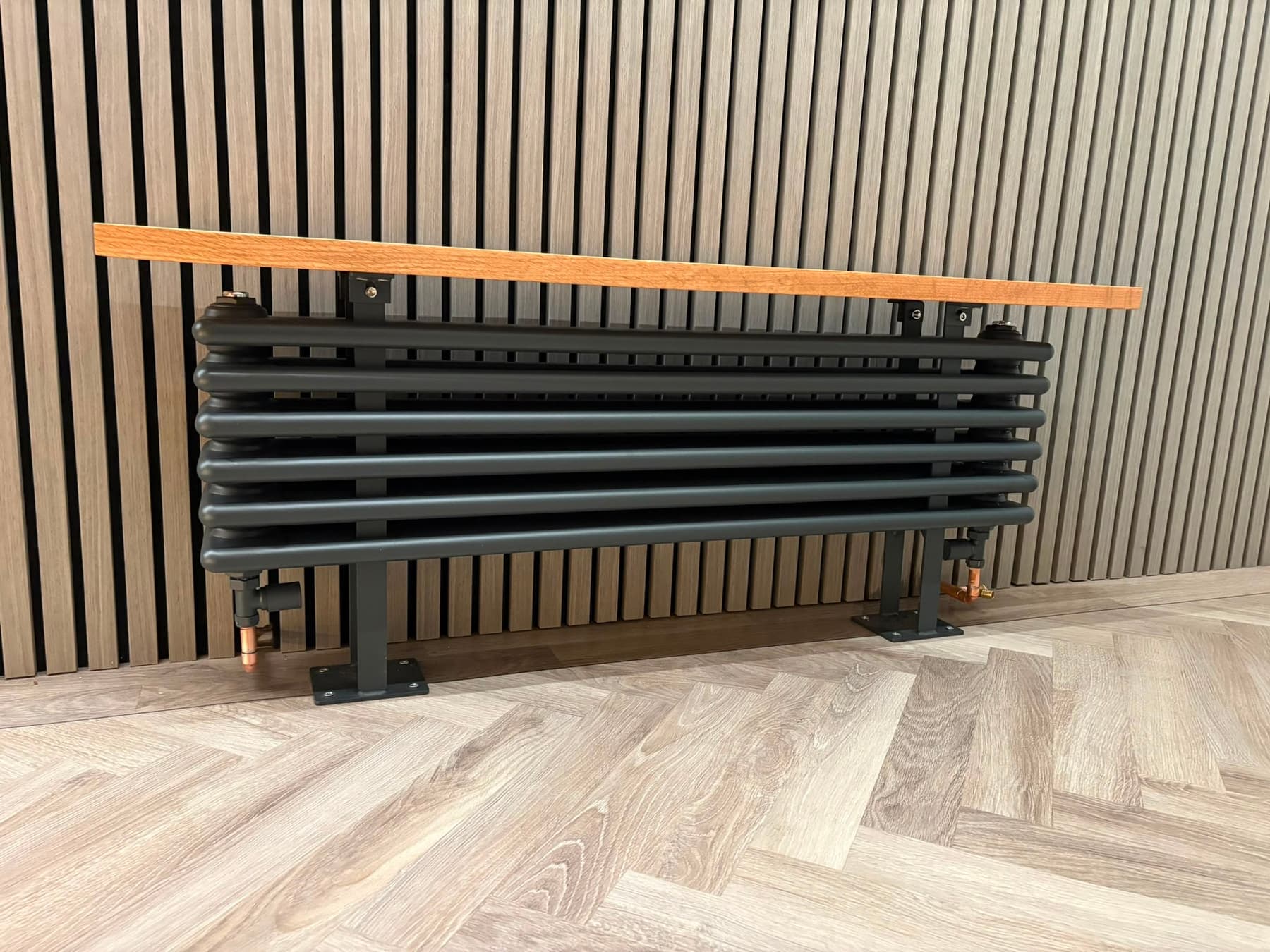 Anthracite designer column radiator with an oak shelf against a slatted wood wall